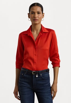 Blusa di raso rossa con colletto a punta, chiusura con bottoni e maniche a tre quarti, abbinata a jeans in denim scuri e una cintura nera.