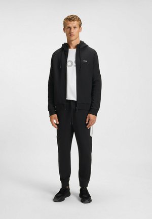 Black zip-up hoodie with drawstrings, paired with matching joggers featuring side stripes, worn over a white logo t-shirt and black sneakers.