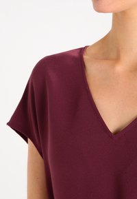 Burgunderfarbener Oberteil aus glattem Stoff, mit V-Ausschnitt und kurzen Ärmeln, lockerer Passform und minimalistischem Design.