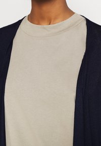 T-shirt beige chiaro con collo rotondo, indossato sotto un cardigan aperto di maglia blu navy, che mette in risalto una texture liscia e un design casual.