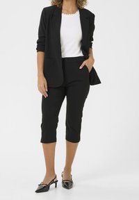 Vrouw in zwarte cropped pants, zwarte blazer met opgerolde mouwen, witte gerimpelde top en zwarte kitten heels, staande voor een witte achtergrond.