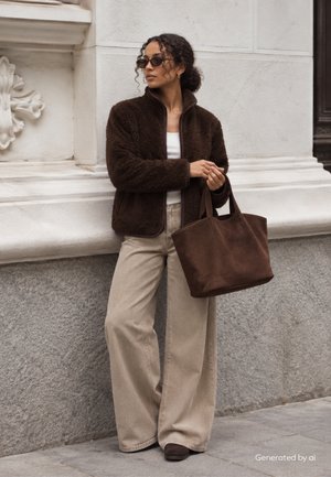 Femme portant une veste duveteuse marron foncé et un pantalon beige à jambes larges, tenant un grand sac cabas marron, debout contre un mur en pierre en extérieur.