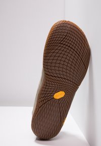 Semelle d'une chaussure beige avec une semelle en caoutchouc marron texturé et un logo Vibram jaune, reposant verticalement contre un mur blanc sur une surface blanche.
