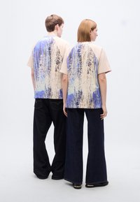 Γραφικό βαμβακερό T-shirt με μπεζ εμπρός μέρος και μπλε αφηρημένο σχέδιο στο πίσω μέρος, συνδυασμένο με παντελόνια από σκούρο τζιν με φαρδιά μπατζάκια.
