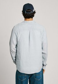 Light blue long-sleeve shirt with a back pleat, soft texture, and rounded hem, worn with blue jeans and a navy cap.