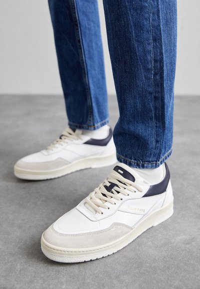 Białe i beżowe sneakersy z granatowymi akcentami, wykonane z fakturowanej zamszowej i skórzanej materiału, okrągły czubek, płaska podeszwa i sznurowane zapięcie. Noszone z niebieskimi dżinsami.
