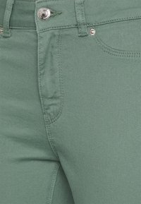 Vihreät denimhousut, joissa on sileä pinta ja klassinen viiden taskun malli, hopeanväriset metalliosat ja vetoketjukiinnitys.