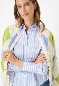 Cardigan in maglia color crema con motivi geometrici verdi e blu; davanti con bottoni. Camicia a righe azzurre e bianche con colletto, indossata sotto.
