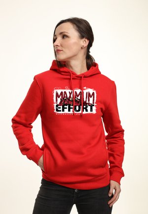 Femme portant un sweat à capuche rouge avec l’inscription "MAXIMUM EFFORT" et des personnages de Deadpool sur le devant, debout les mains dans les poches.