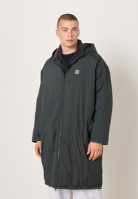 Mladý muž v dlhom tmavom zimnom kabáte s kapucňou, predným zipsom a nášivkou s logom, stojaci pred jednoduchým svetlým pozadím.