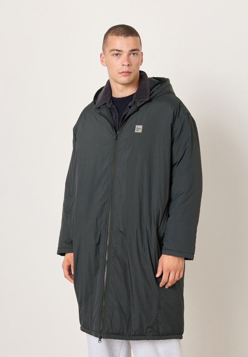 Mladý muž v dlhom tmavom zimnom kabáte s kapucňou, predným zipsom a nášivkou s logom, stojaci pred jednoduchým svetlým pozadím.