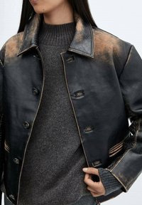 Vrouw met een zwarte versleten leren jas over een donkergrijze geribbelde gebreide trui, die met één hand de revers van de jas vasthoudt.