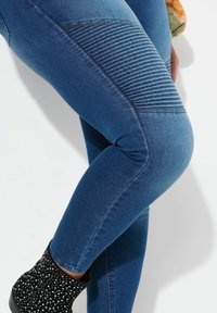 Leggings de mezclilla azul con textura acanalada en el muslo, ajuste slim, y una bota negra con manchas blancas en la parte inferior de la imagen.