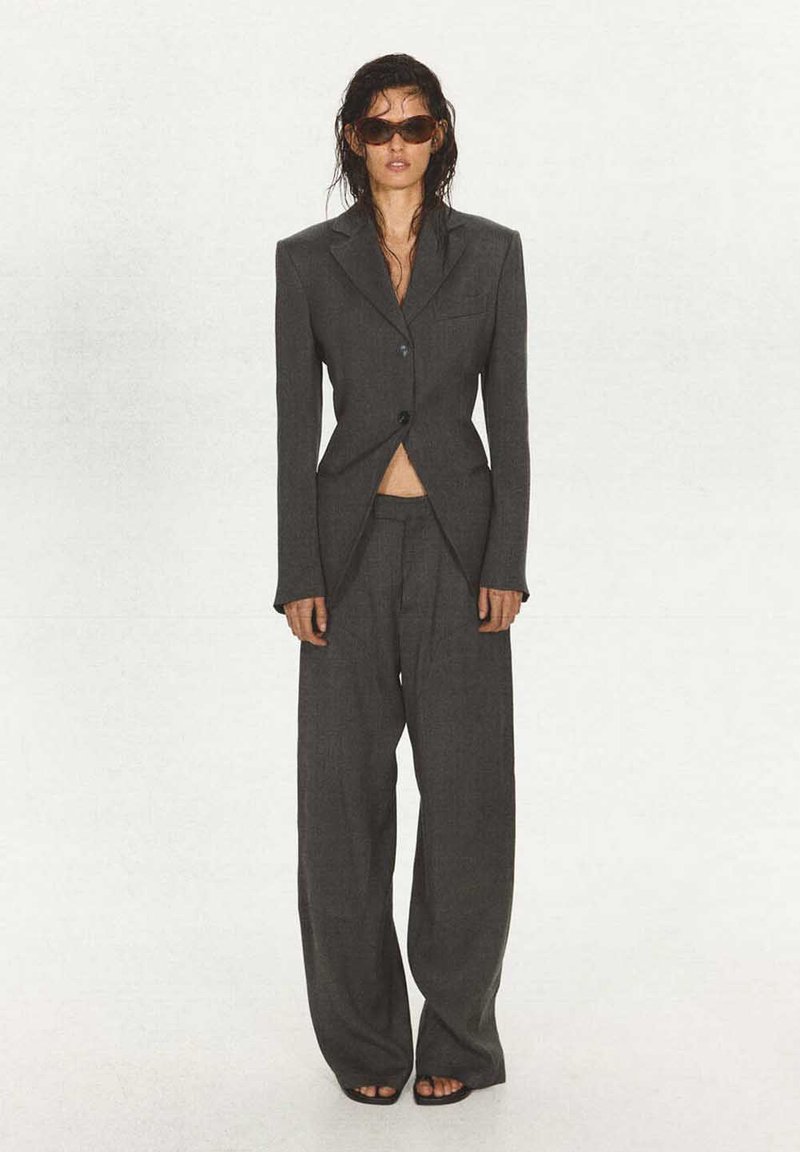 Femme aux cheveux mouillés portant un costume sombre surdimensionné avec un pantalon large et des lunettes de soleil, debout devant un fond clair uni.
