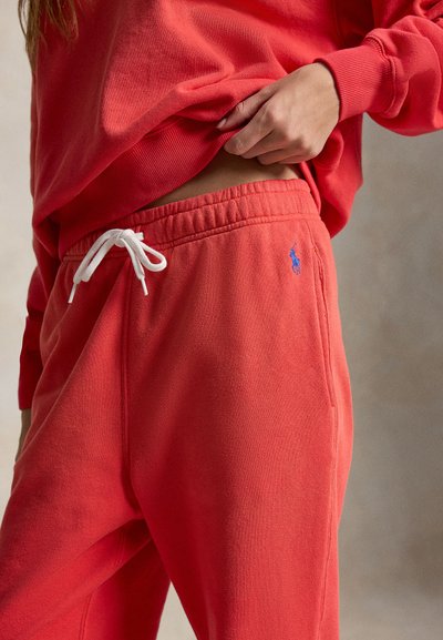 Osoba poprawia dolną krawędź czerwonej bluzy, nosząc dopasowane czerwone spodnie dresowe z białym sznurkiem i małym niebieskim haftowanym logo po boku.