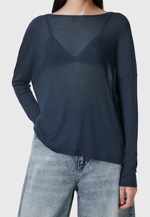 Kvinde iført en gennemsigtig navyblå langærmet top over en mørk bh kombineret med lyseblå løstsiddende jeans, stående med den ene hånd i lommen.