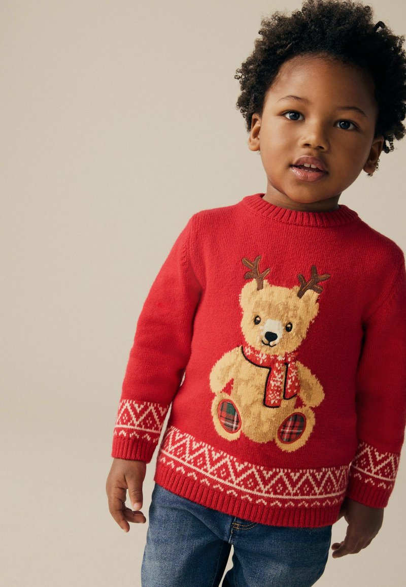 Kleines Kind trägt einen roten Pullover mit einem Teddy, der Rentiergeweih und einen Schal trägt, kombiniert mit blauen Jeans, vor einem schlichten Hintergrund stehend.