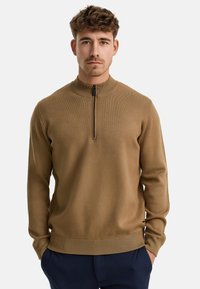 Brauner Pullover mit hohem Kragen und einem Viertel-Reißverschluss, gerippter Textur und enganliegenden Bündchen, aus einem weichen Strickmaterial gefertigt, über einer navyblauen Hose getragen.