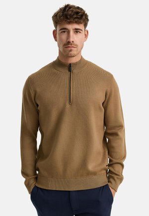 Brun højhalset pullover med en kvart lynlås, ribbet tekstur og tætsiddende manchetter, lavet af et blødt strikmateriale, båret over marineblå bukser.