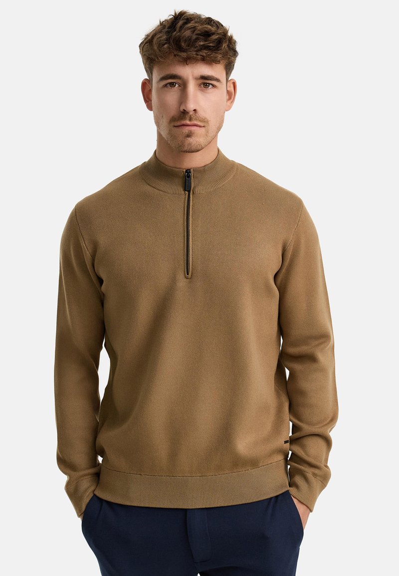 Brauner Pullover mit hohem Kragen und einem Viertel-Reißverschluss, gerippter Textur und enganliegenden Bündchen, aus einem weichen Strickmaterial gefertigt, über einer navyblauen Hose getragen.