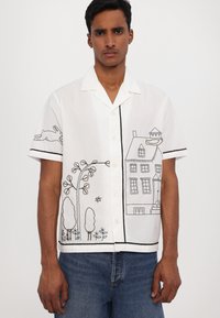 Camisa blanca de manga corta con diseños bordados en negro de una casa, árboles y un conejo. Presenta un cuello y cierre de botones.