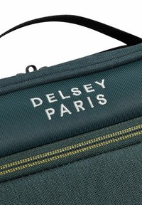 Bolso de tela verde oscuro con un asa superior, un logo blanco de "DELPSEY PARIS" y costuras amarillas a lo largo de la cremallera. Superficie texturizada.