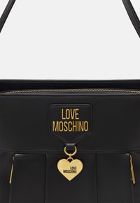 Bolso negro plisado con herrajes dorados, con una etiqueta con el logo "LOVE MOSCHINO" y un colgante en forma de corazón. Textura de cuero suave.