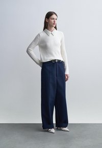 Egy nő fehér bordázott pulóvert visel gyöngygallérral, tengerészkék bő szárú nadrágot, fekete övet és fehér hegyes orrú cipőt, miközben szürke padlón áll.