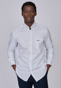 Chemise blanche à boutons avec des pois noirs, dotée d'un col pointu et d'une poche. Manches longues avec des poignets boutonnés.