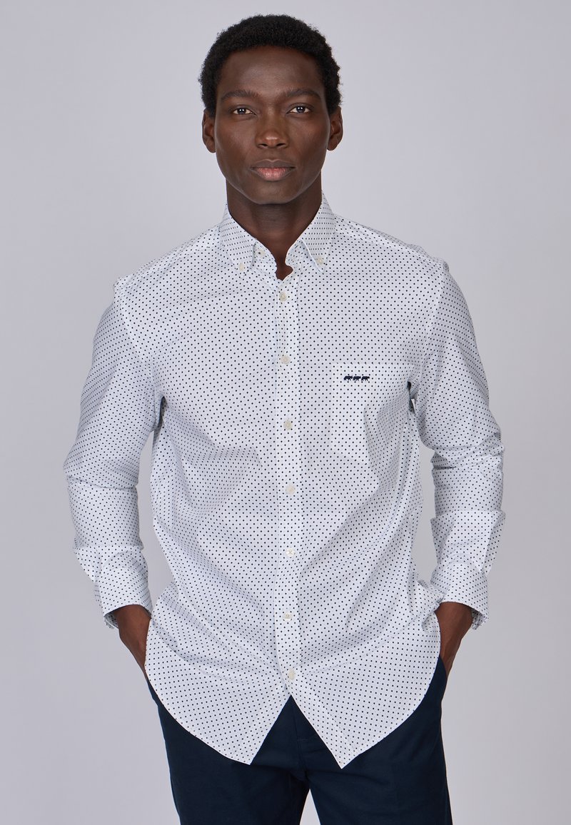 Chemise blanche à boutons avec des pois noirs, dotée d'un col pointu et d'une poche. Manches longues avec des poignets boutonnés.