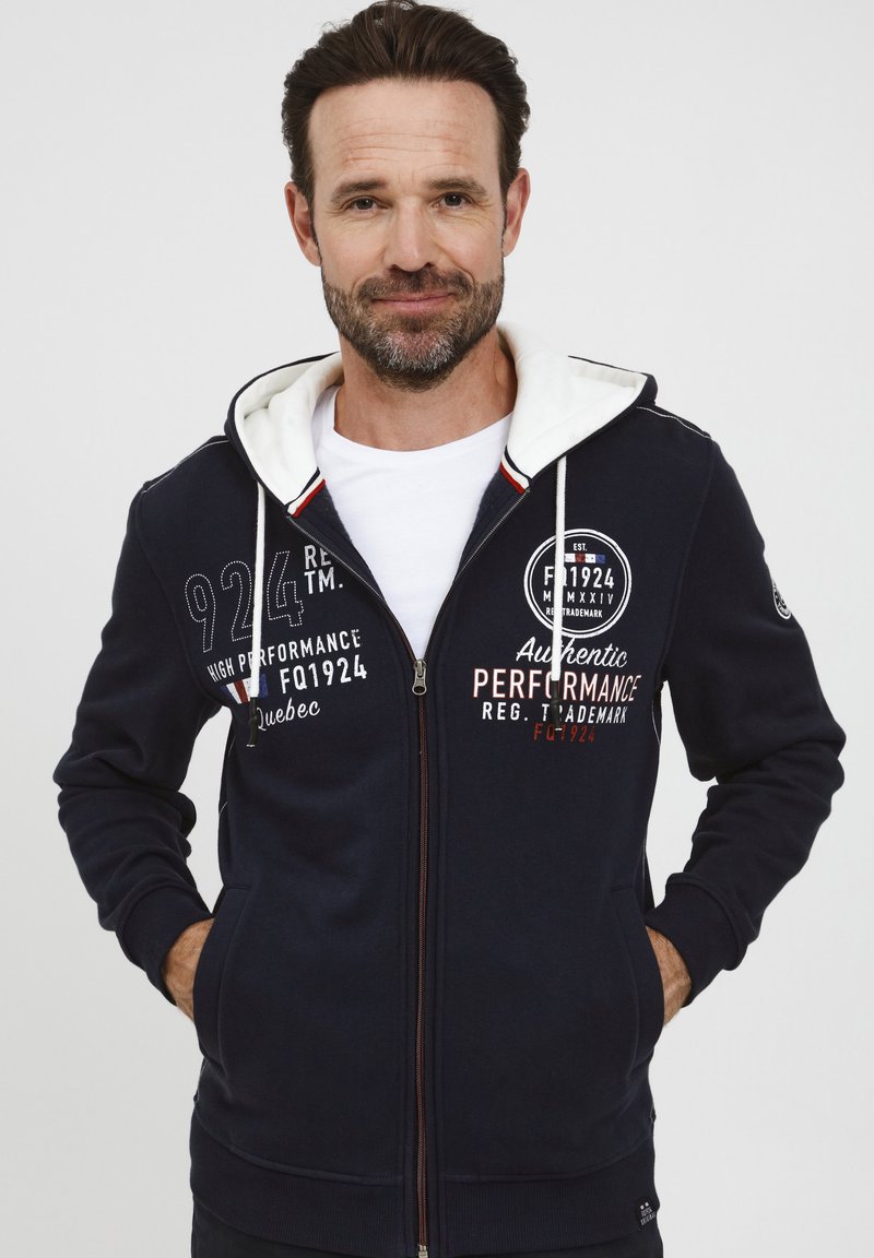 Felpa con zip color navy e fodera bianca nella cappuccio, caratterizzata da stampe del logo e testi in bianco e rosso. Tessuto morbido con vestibilità rilassata.