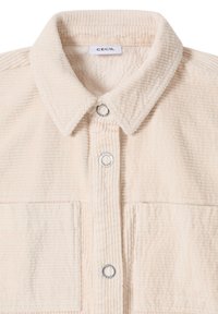 Beige corduroy-skjorte med spids krave, to påsyede lommer og sølvfarvede trykknapper. Tekstureret stof med lodret ribbing.