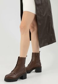 Stivali alla caviglia in pelle marrone con lacci sul davanti, zip laterale, tacco chunky e suola testurizzata, abbinati a un lungo cappotto marrone.