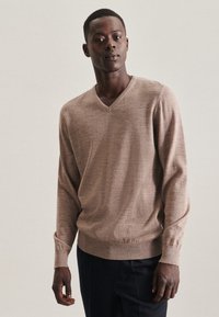V-Ausschnitt-Pullover in einem hellbraunen Farbton, aus strukturiertem Stoff gefertigt, mit gerippten Bündchen und Saum. Das Modell posiert mit einer leicht erhobenen Hand.