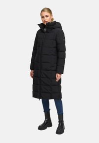 Abrigo acolchado negro con capucha, con un diseño largo y rectangular, patrón acolchado horizontal y cremallera frontal. Combinado con jeans oscuros y botas.