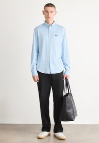 Lys blå langermet skjorte med krage, svarte bukser, og en svart lær toalettveske. Modellen har på seg hvite joggesko.