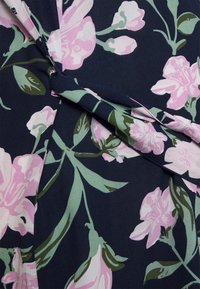 O tecido do vestido floral apresenta flores rosas e verdes sobre um fundo azul-marinho, com pregas e seções sobrepostas que realçam a textura e o design.