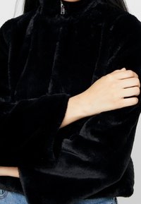 Sudadera de piel sintética negra con cuello alto y detalle de cremallera. Textura suave, mangas anchas y diseño ligeramente recortado.