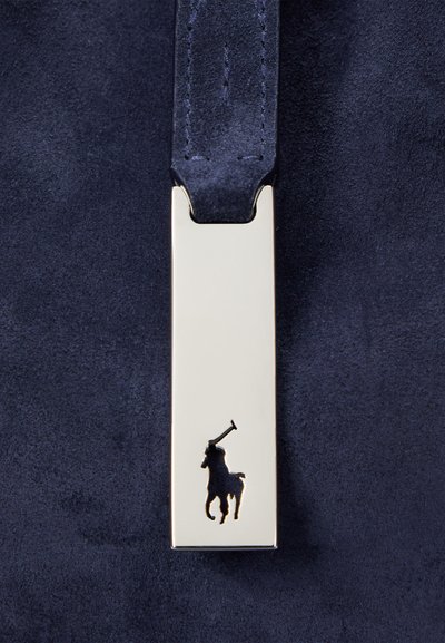 Detalj av marinblå mockaväska med en slät guldtonad metallbit och en svart siluett av en polospelare. Elegant och modern design.