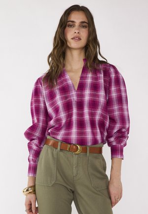 Femme portant un chemisier à carreaux rose et blanc avec des manches bouffantes, rentré dans un pantalon vert olive avec une ceinture en cuir marron et un bracelet en or.