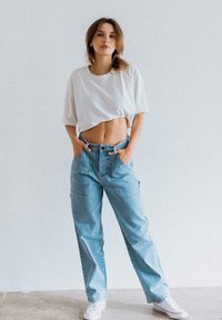 Helle blaue Jeans-Cargo-Hose mit großen Fronttaschen, kombiniert mit einem weißen, kurzen T-Shirt. Das Outfit wird mit weißen Turnschuhen abgerundet.