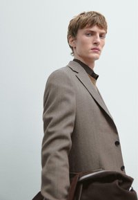 Brązowa marynarka w kratę z klapami z wycięciem i czarnymi guzikami, noszona na ciemnej koszuli. Osoba trzyma brązową torbę, ma krótkie jasne włosy.