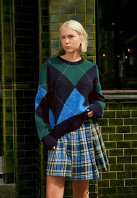 Pull en tricot surdimensionné avec un motif géométrique en vert, bleu et marine, associé à une jupe plissée en tartan dans des tons de bleu et de jaune.