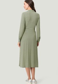 Langsleeve midi-jurk in lichtgroen met een geometrisch zwart en geel patroon. Glad stof met een getailleerde taille en een uitlopend rokje.