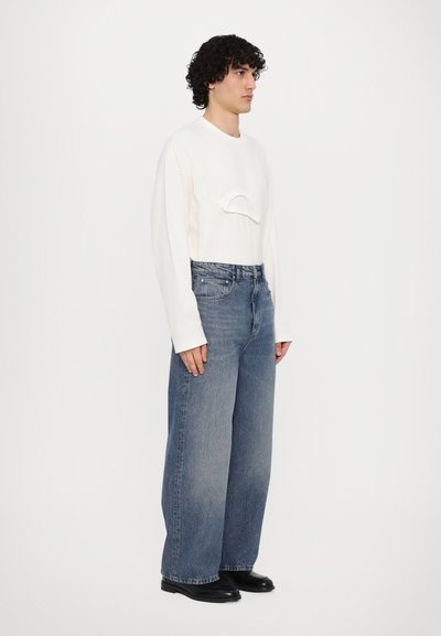 Weißes Langarmhemd mit einer curved Detail auf der Brust, kombiniert mit weit geschnittenen blauen Denim-Jeans und schwarzen Stiefeletten.
