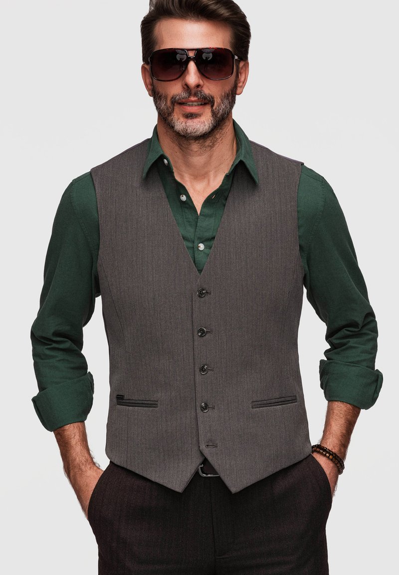 Homme avec une barbe portant une chemise vert foncé, un gilet gris boutonné, des lunettes de soleil et des bracelets, debout les mains dans les poches.