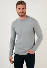 Maglione grigio chiaro a trama, con scollatura rotonda, polsini e orlo a costine, indossato sopra jeans scuri. Motivo esagonale sottile su tutta la superficie.