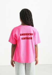 Różowy bawełniany t-shirt z krótkim rękawem, z grubym czerwonym napisem "AMERICAN VINTAGE" na plecach. Noszony z szarymi spodniami dresowymi.