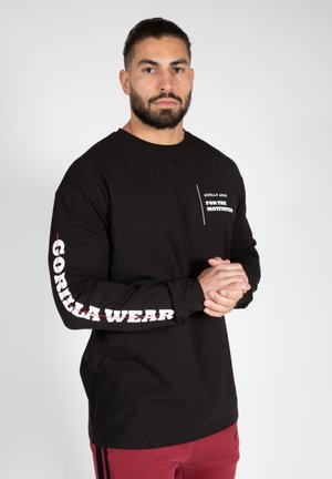 Sort langærmet skjorte lavet af bomuld, med hvid tekst "GORILLA WEAR" på ærmet og et brystlogo. Bæret med røde bukser.