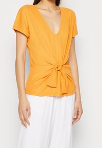 Top naranja texturizado con escote en V y mangas cortas, con un diseño de frente anudado; combinado con parte inferior blanca de corte holgado.